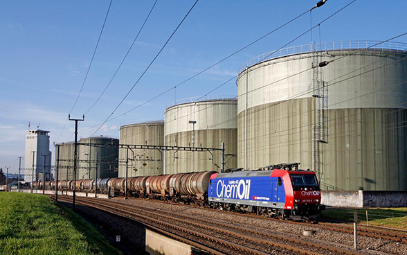 Eine Güterzuglok mit Kesselwagen fährt an vier grossen runden Tanks vorbei.